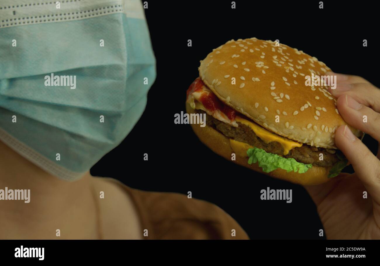 Petrozavodsk. Russia. June 23. 2020: Female hand holding a burger and a blue glass at the mouth of a closed medical mask on a black background Stock Photo