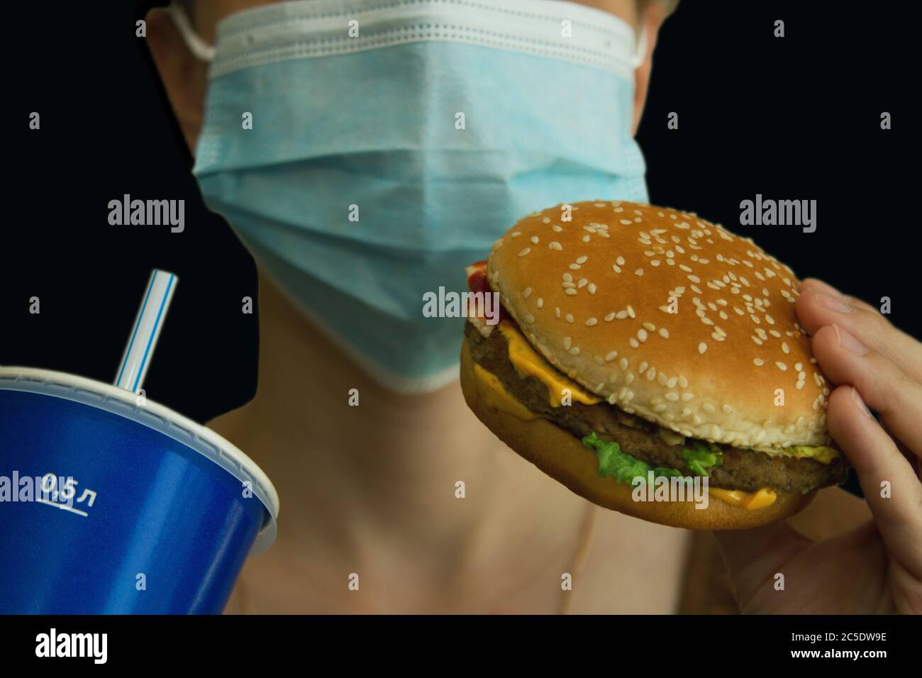 Petrozavodsk. Russia. June 23. 2020: Female hand holds burger to mouth with closed medical mask on a black background Stock Photo