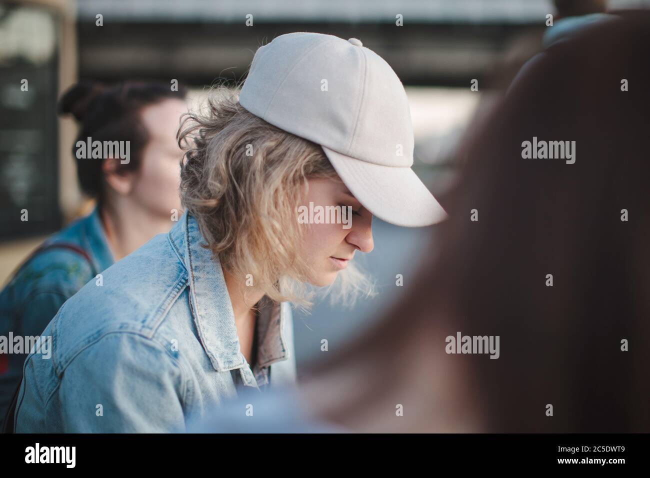 Beautiful blonde girl in a cap with short hair sits in a group of girls, looks down. Loneliness among friends. Selective focus. Stock Photo