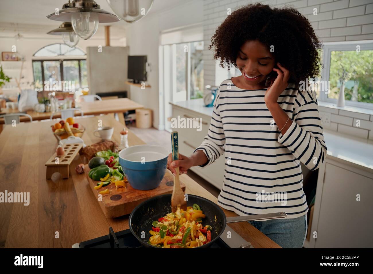 Happy african young woman talking on smartphone while preparing meal during covid-19 lockdown Stock Photo