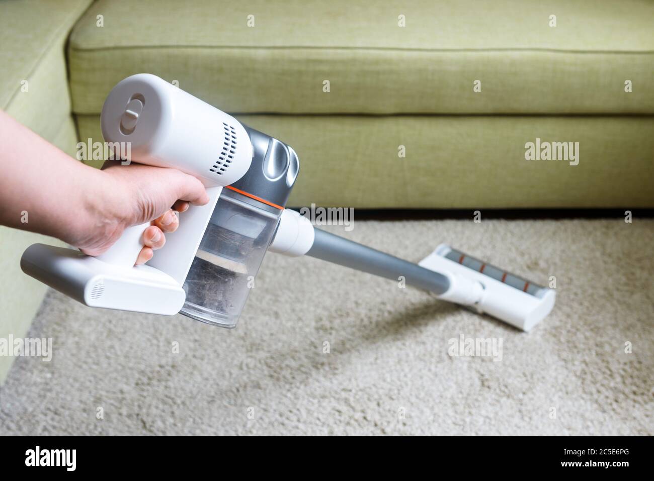 Wireless vacuum cleaner used on carpet in room. Housework with new upright hoover. Person holds modern white vacuum cleaner by sofa. Home cleaning, ca Stock Photo