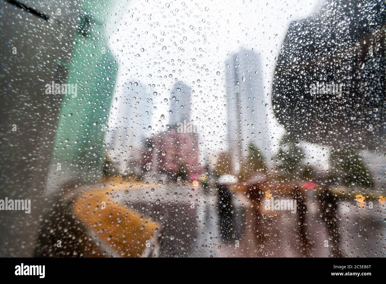 Rain droplets on a window with modern city architecture and people carrying umbrellas out-of-focus in the background. Stock Photo