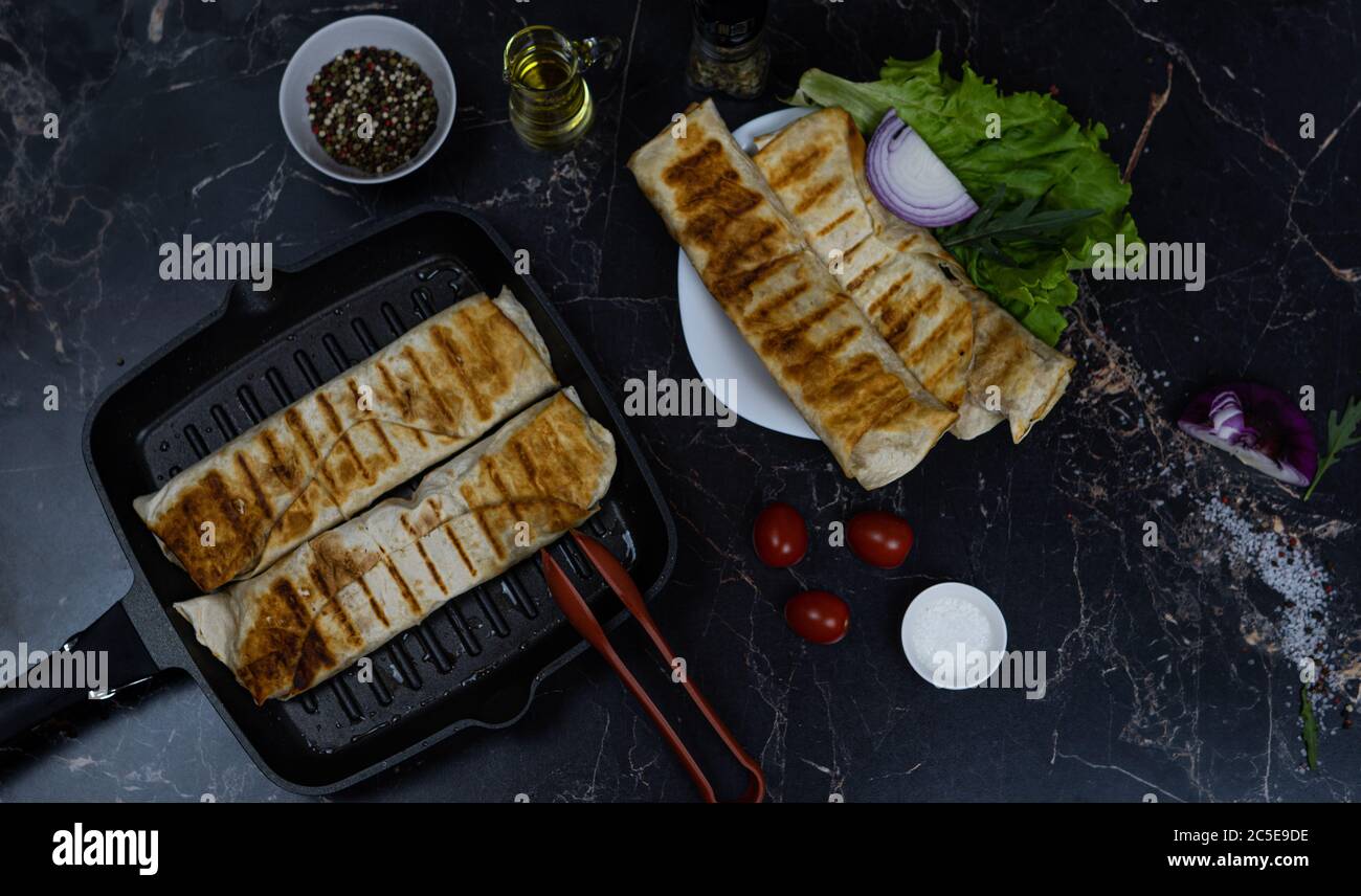 Grilled chicken Shawarma with spices and vegetables. Shawarma on a grill pan. Shawarma on a dark background. Stock Photo
