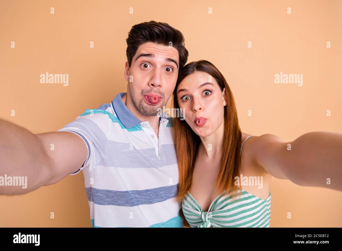 Photo of positive crazy foolish hipster couple make selfie enjoy honeymoon  show tongue out wear striped t-shirt dress skirt isolated over pastel color  Stock Photo - Alamy