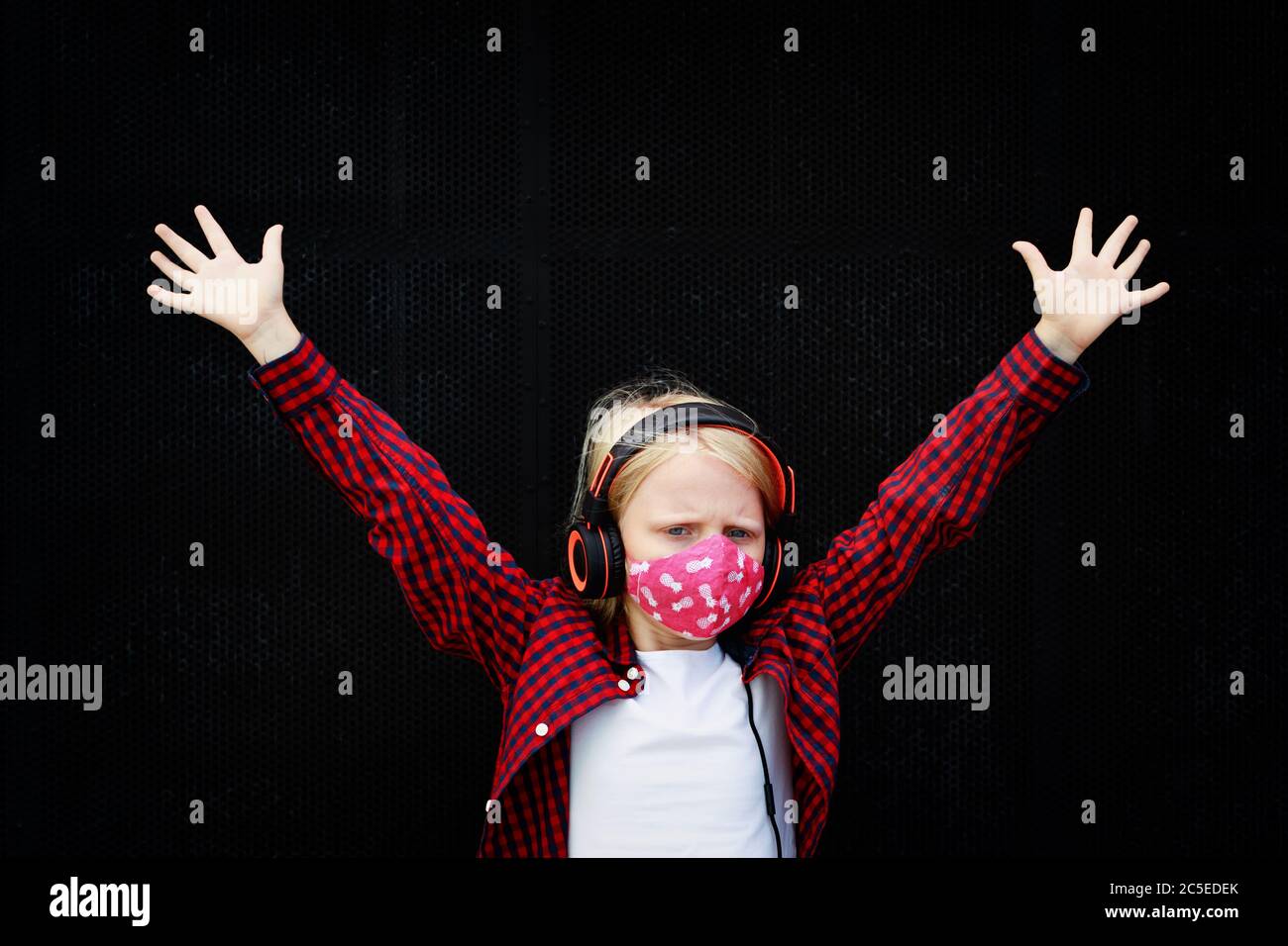 Young child in headphones listening music with fun raise hands up. Funny girl wear stylish protective face mask due coronavirus COVID 19 pandemic Stock Photo