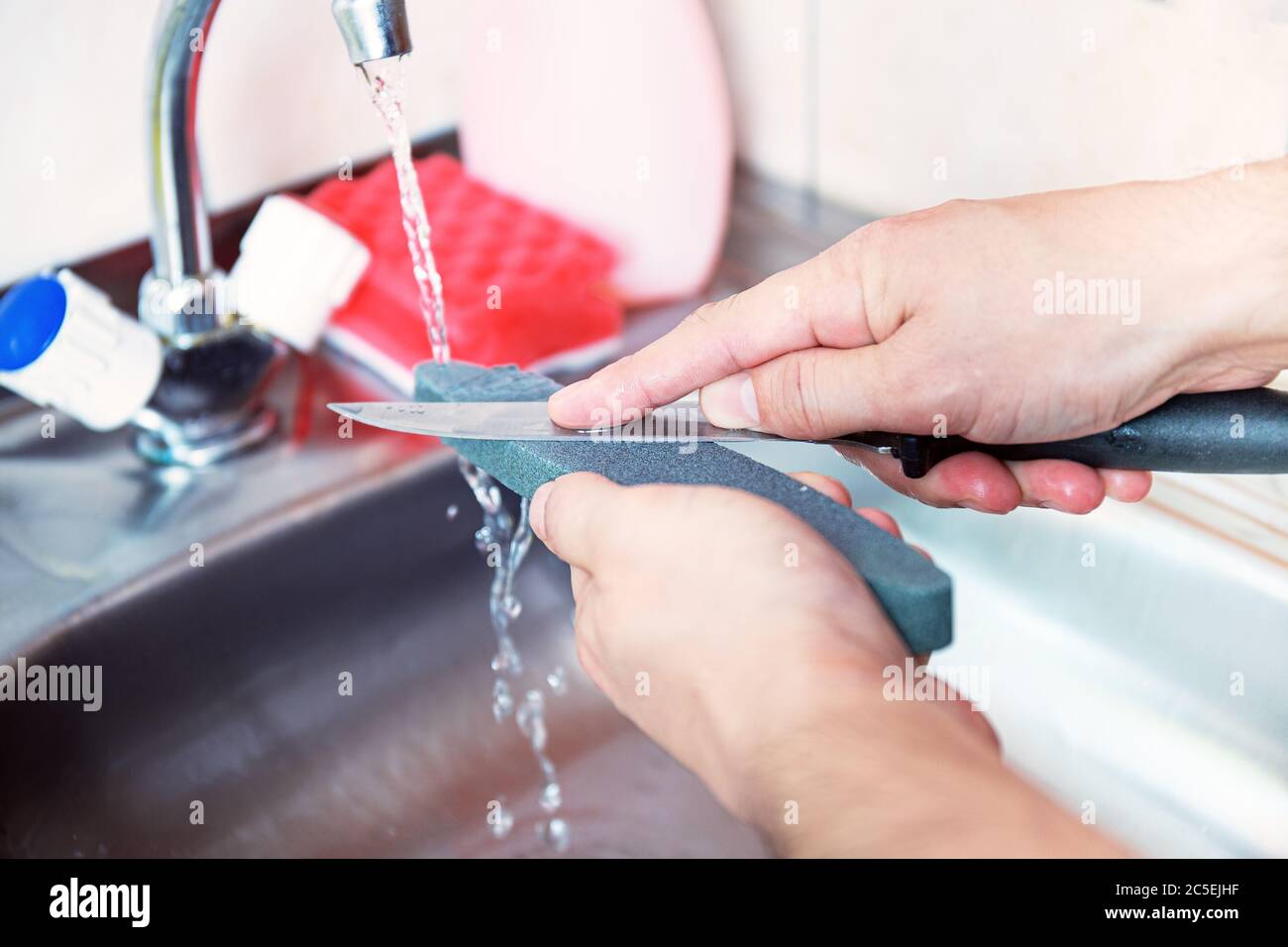 Close-up hands of a man carefully manual sharpen a knife under a stream of water on a grindstone. Home household sharpening. Care for kitchen cutlery. Stock Photo