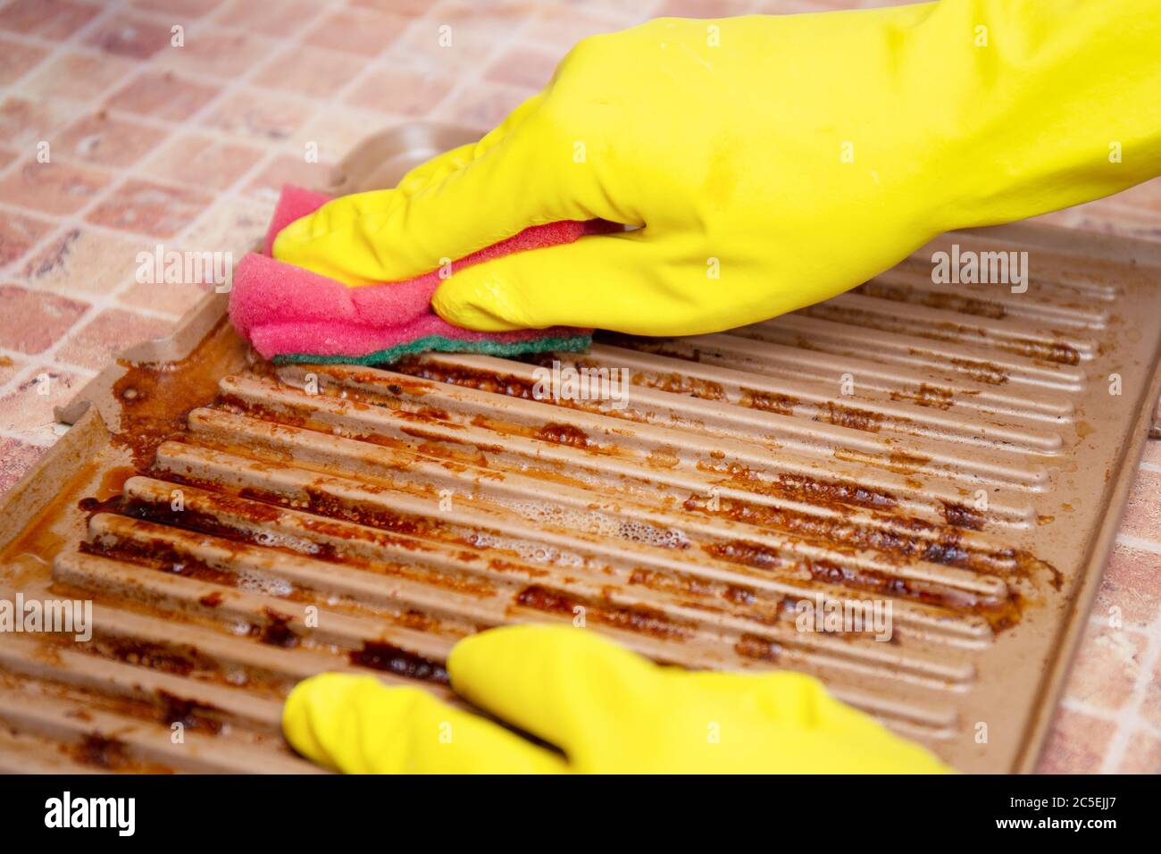 Hands in yellow household gloves remove grease from a dirty electric grill using a sponge and a special spray. Drops of fat after frying. Care for the Stock Photo