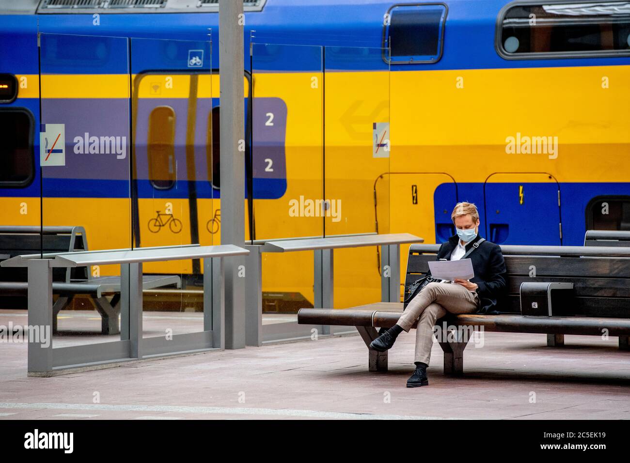 A man wearing a facemask is seen seated outside the train while reading papers at a train station.Residents of Rotterdam can take the train starting 1 July with mandatory wearing of face masks. In addition, all seats can be used on buses, trams, metros and trains. The Dutch Railways (NS) will however shrink considerably in the next five years due to the coronavirus crisis as the transport company expects to cut 2,300 jobs. Stock Photo