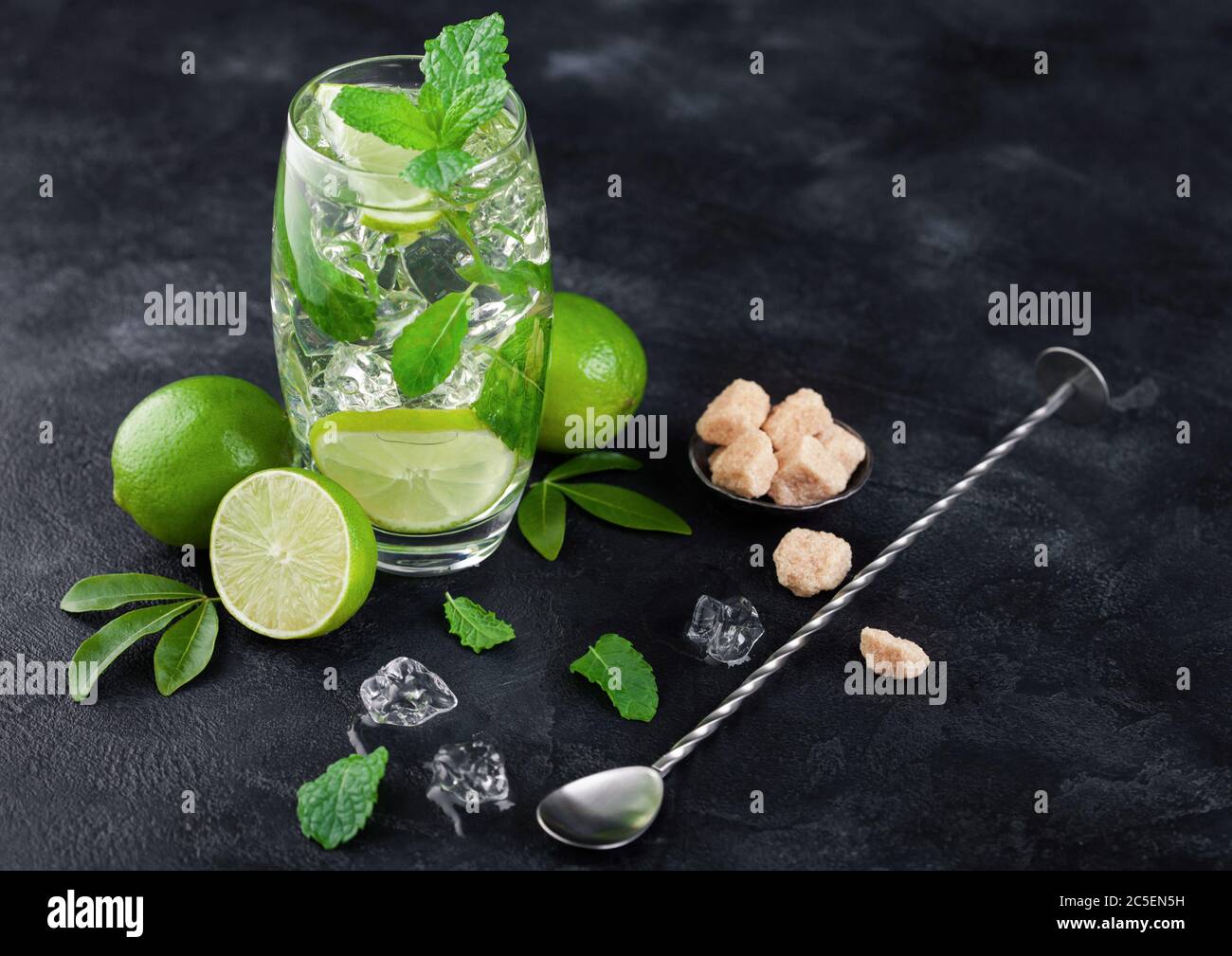 Mojito refreshing summer cocktail in glass with ice cubes mint and lime on black board with spoon and fresh limes with cane sugar. Stock Photo