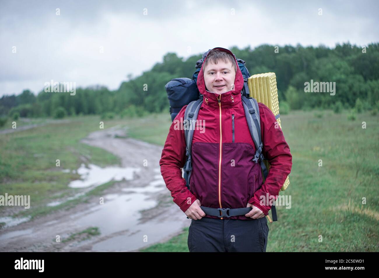 Guy tourist hiker walks along the dirt road with a backpack in a red jacket in the rain Stock Photo