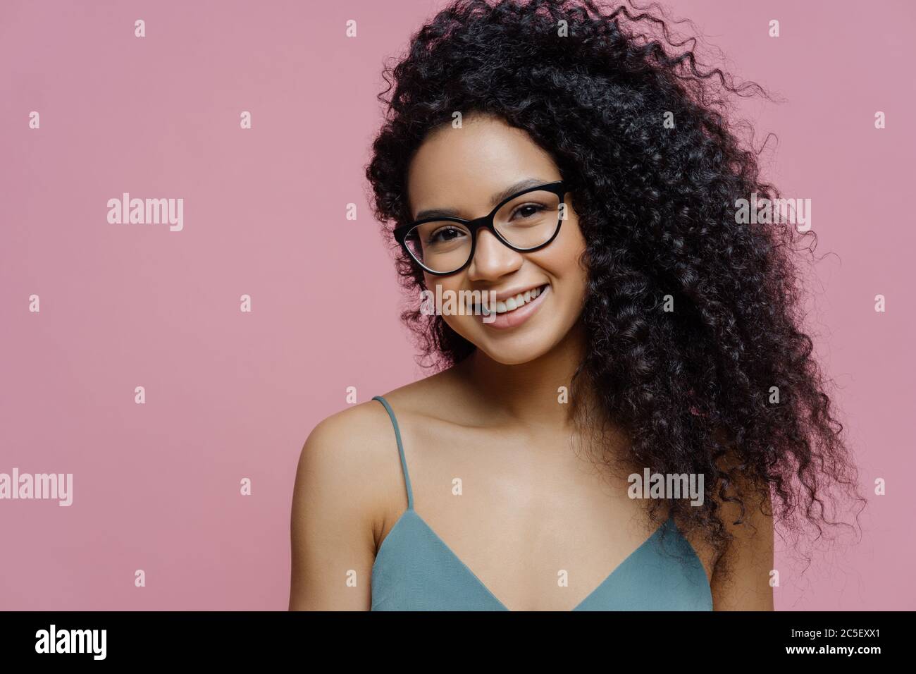 Portrait of attractive dark skinned woman with Afro bushy hair, dressed casually, looks through transparent glasses, has happy smile on face, isolated Stock Photo