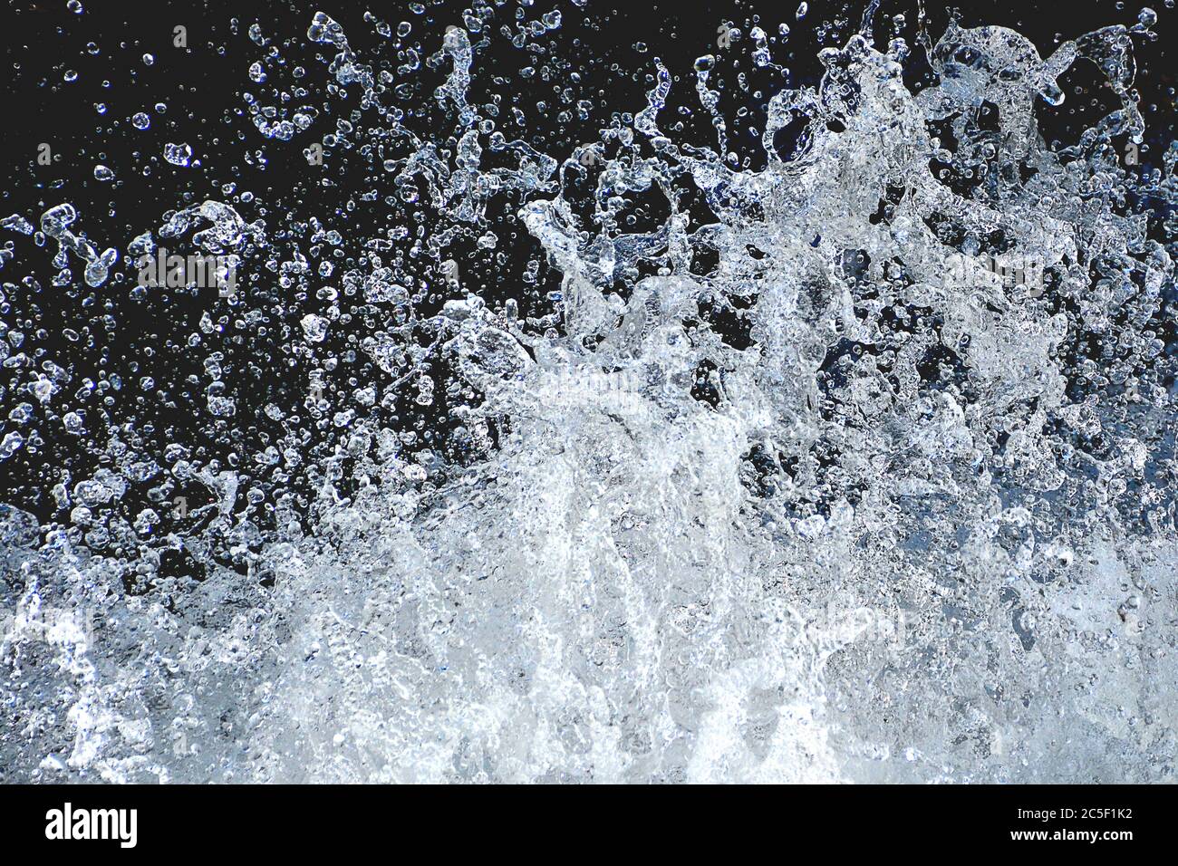 Attention-grabbing closeup of shimmering drops of water splashing up from rushing stream, captured in midair, and isolated against dark background. Stock Photo