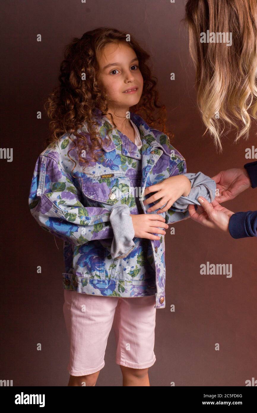 mom helps her daughter get dressed. A girl with long curly hair in white shorts and a colorful jacket, female hands in the frame Stock Photo