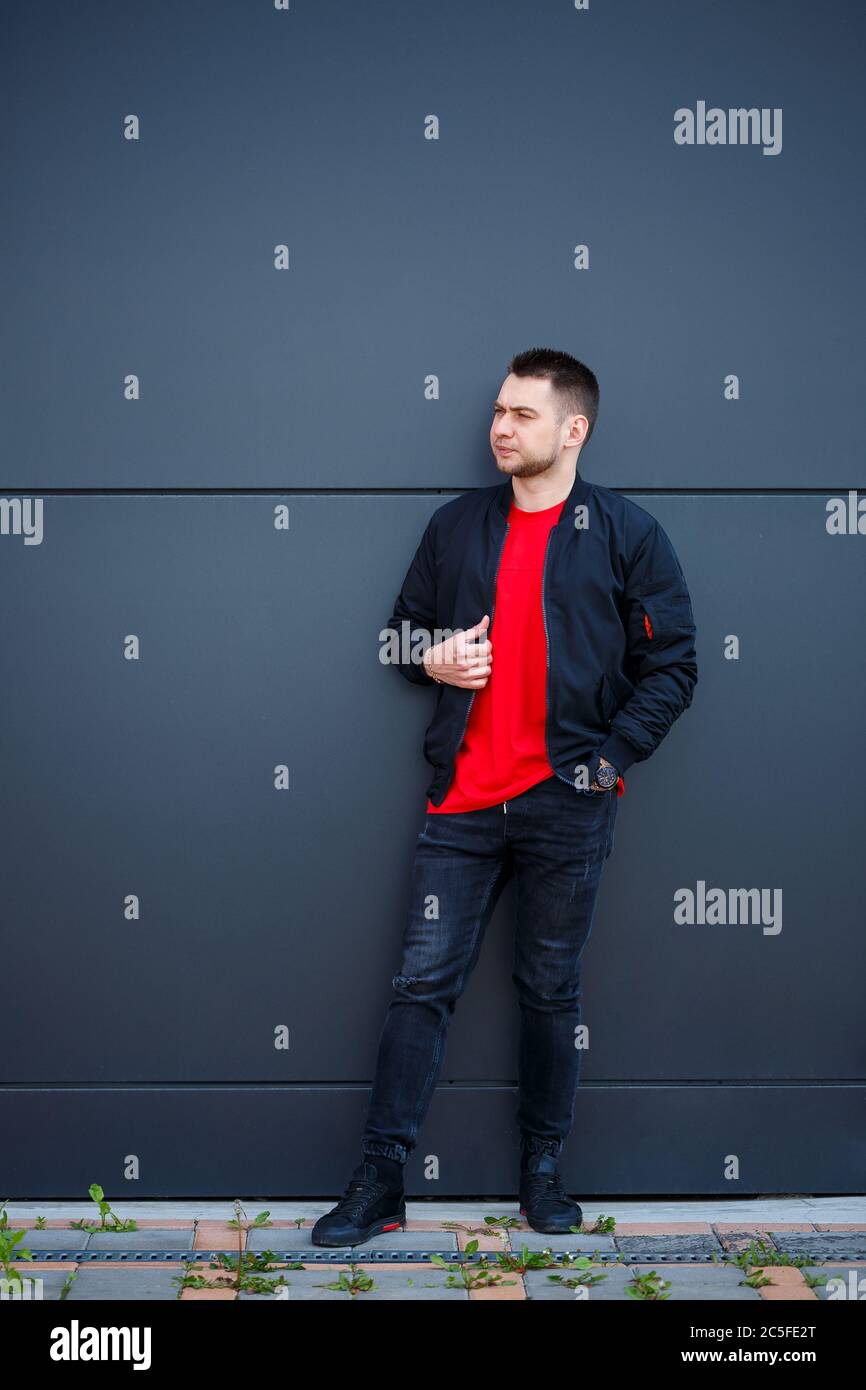 A young man dressed in casual style, a black jacket, jeans, a red T-shirt.  He is against a gray wall Stock Photo - Alamy