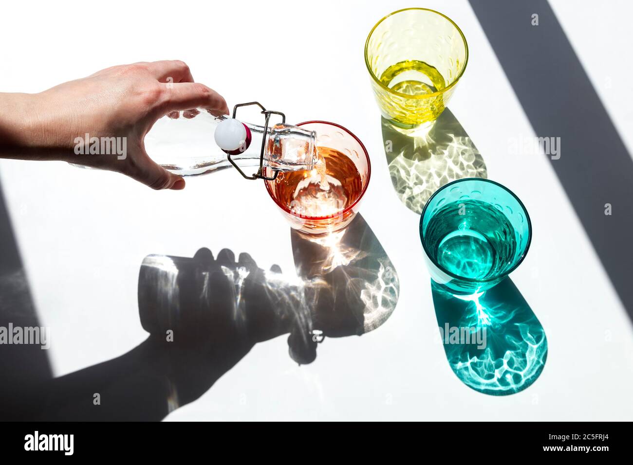 A person pours water into colored glass cups under the strong sunlight that enters through the window and casts its textured, colored shadows on a whi Stock Photo
