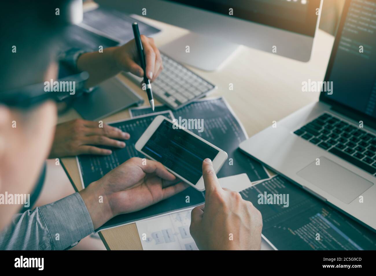Software developers are using smart phones and analyzing together the code written in the program on the computer. Stock Photo