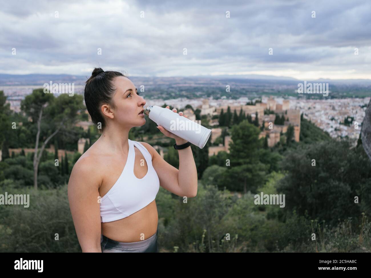 Fit pretty woman drinking water from a reusable bottle after training above the city with the Alhambra in the background at sunset in summer. Stock Photo
