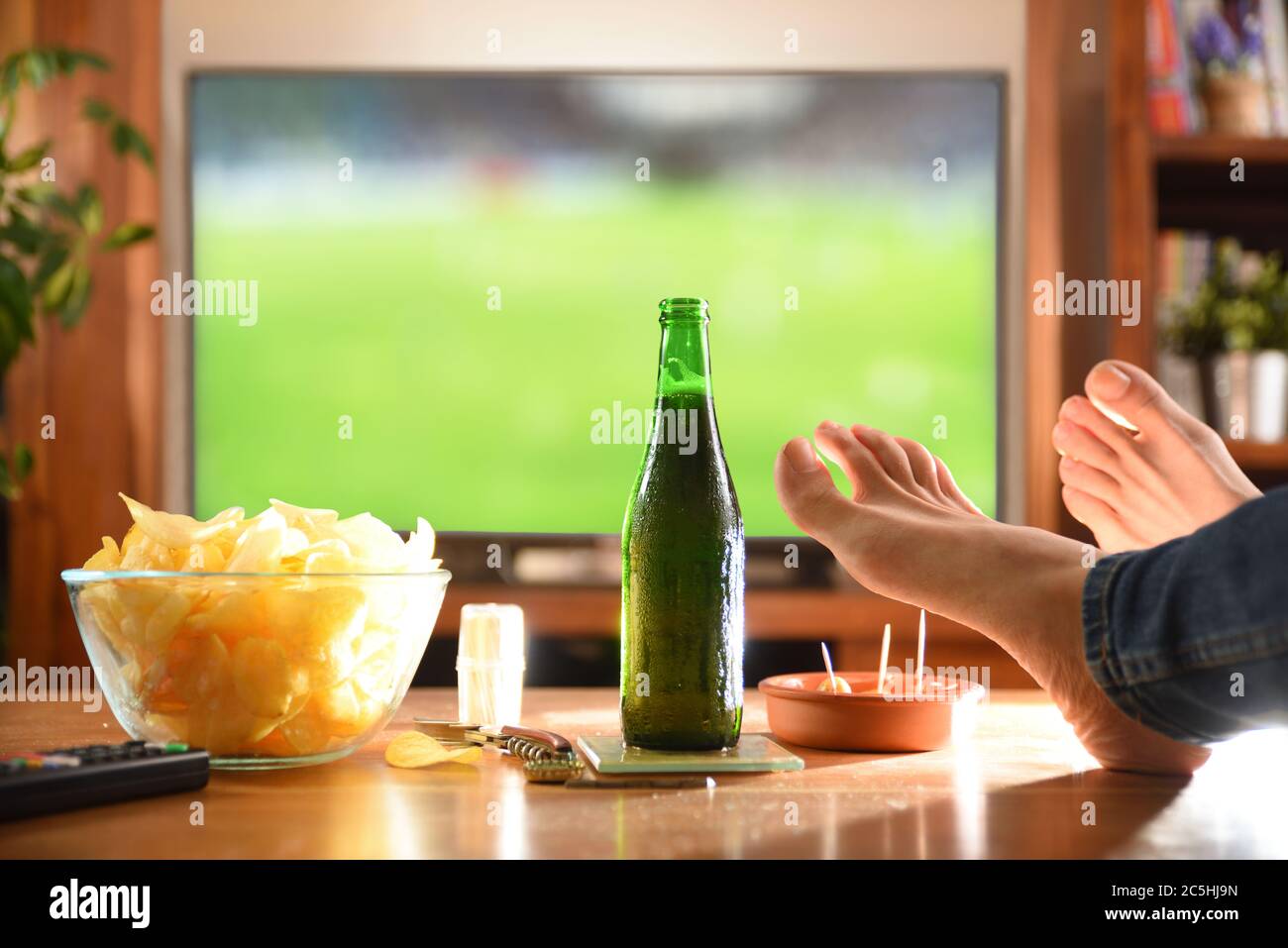 A person's hand holding a brown beer bottle, whil watching a soccer match  on tv. His legs are placed on the wooden table to relax. Stock Photo
