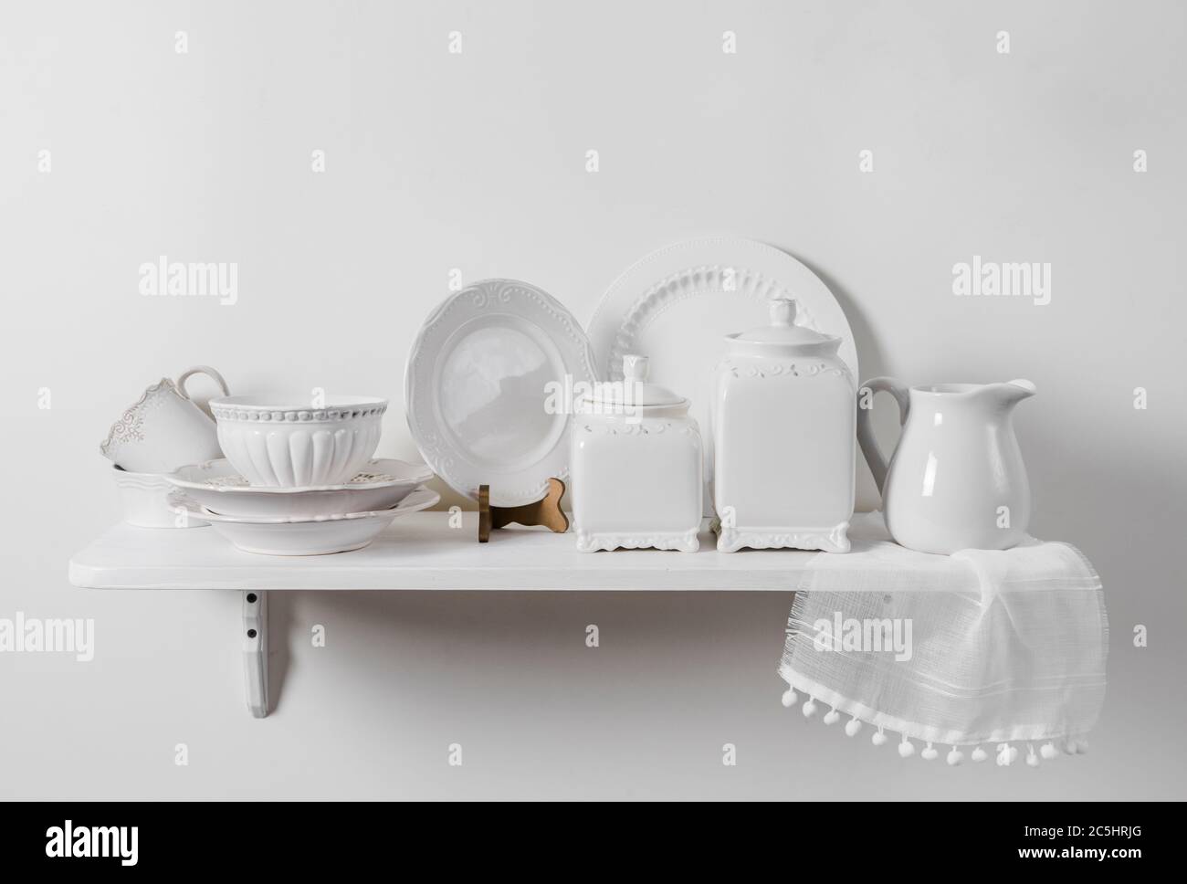 Composition of white wood hanging shelf on wall with country home tableware set on display. Romantic pattern plates, ceramic storage box containers. Stock Photo