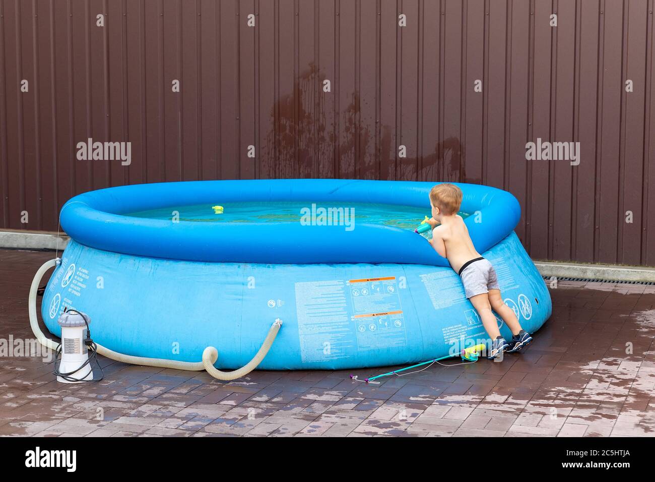 Cute little adorable caucasian blond toddler boy looking into inflatable blue pool enjoy playing with toy fishing rod at home yard on hot summer day Stock Photo