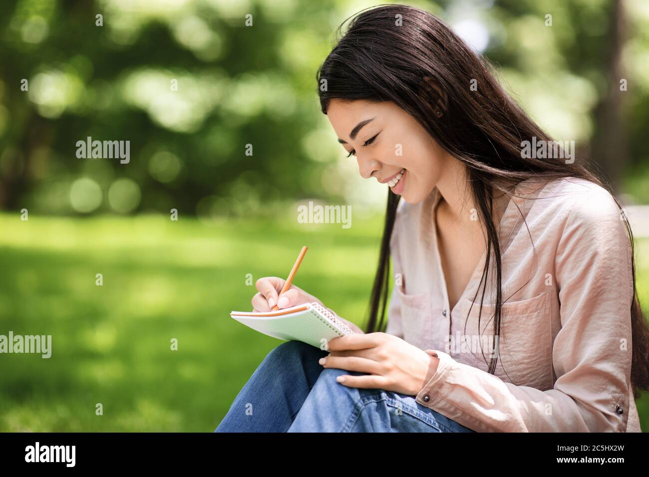 Young asian student girl sitting on lawn in park and writing notes ...