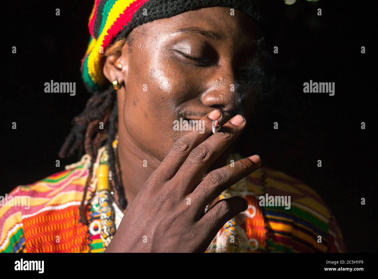 Face of happy young African Rastafarian man smoking outdoors at night ...
