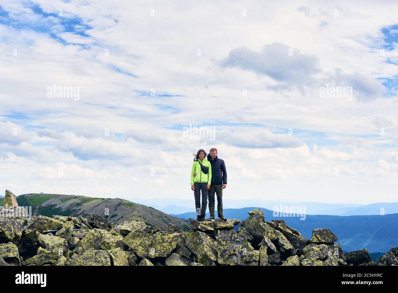 Front view of loving couple hugging, posing and looking at camera, beautiful mountains scenery on background. Mountain hiking, tourists reaching peak together. Wild nature, sport tourism concept. Stock Photo