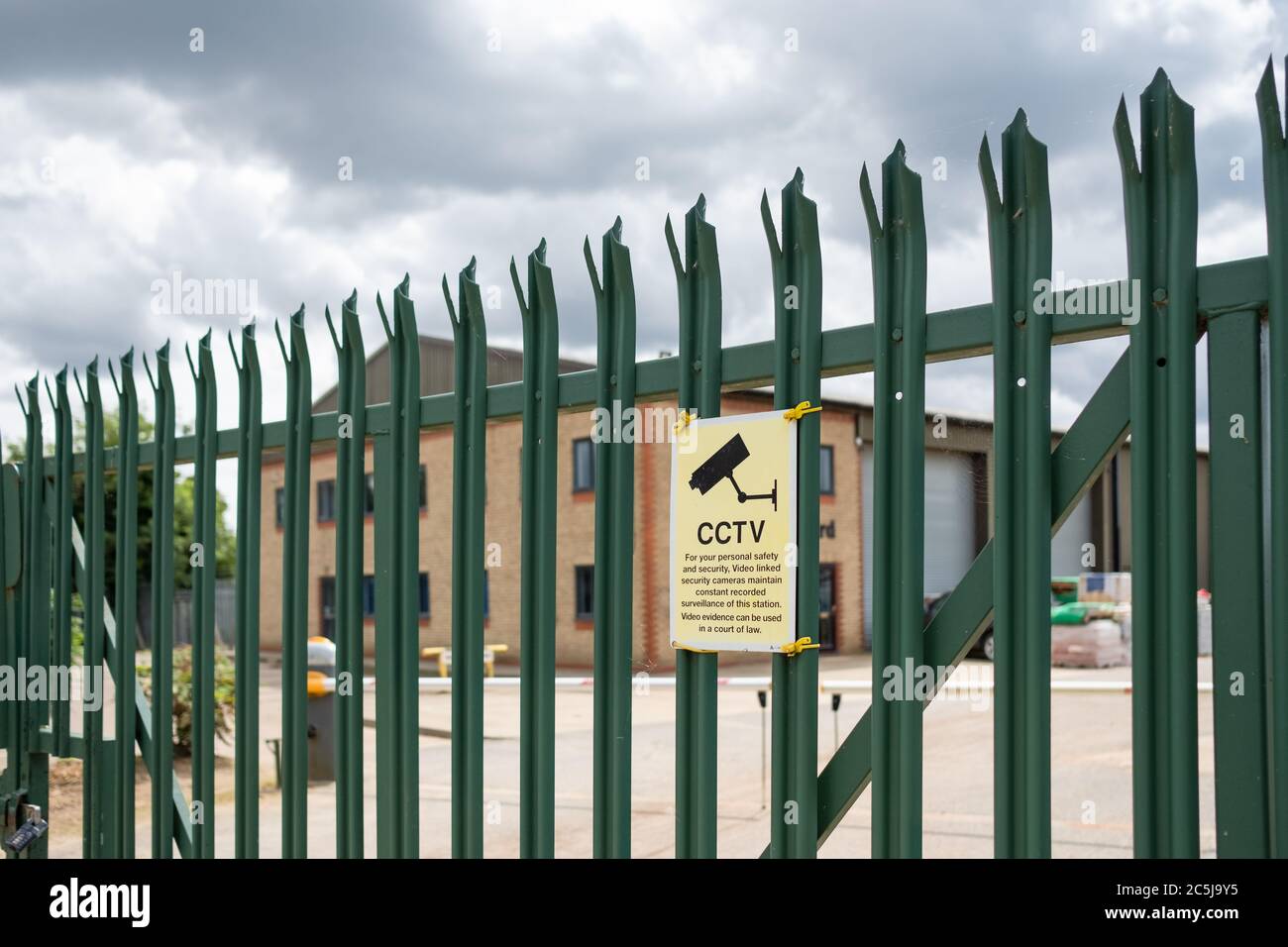Shallow focus of a CCTV security sign seen on a metal, secure industrial fence. Beyond the fence is a large warehouse and office buildings. Stock Photo