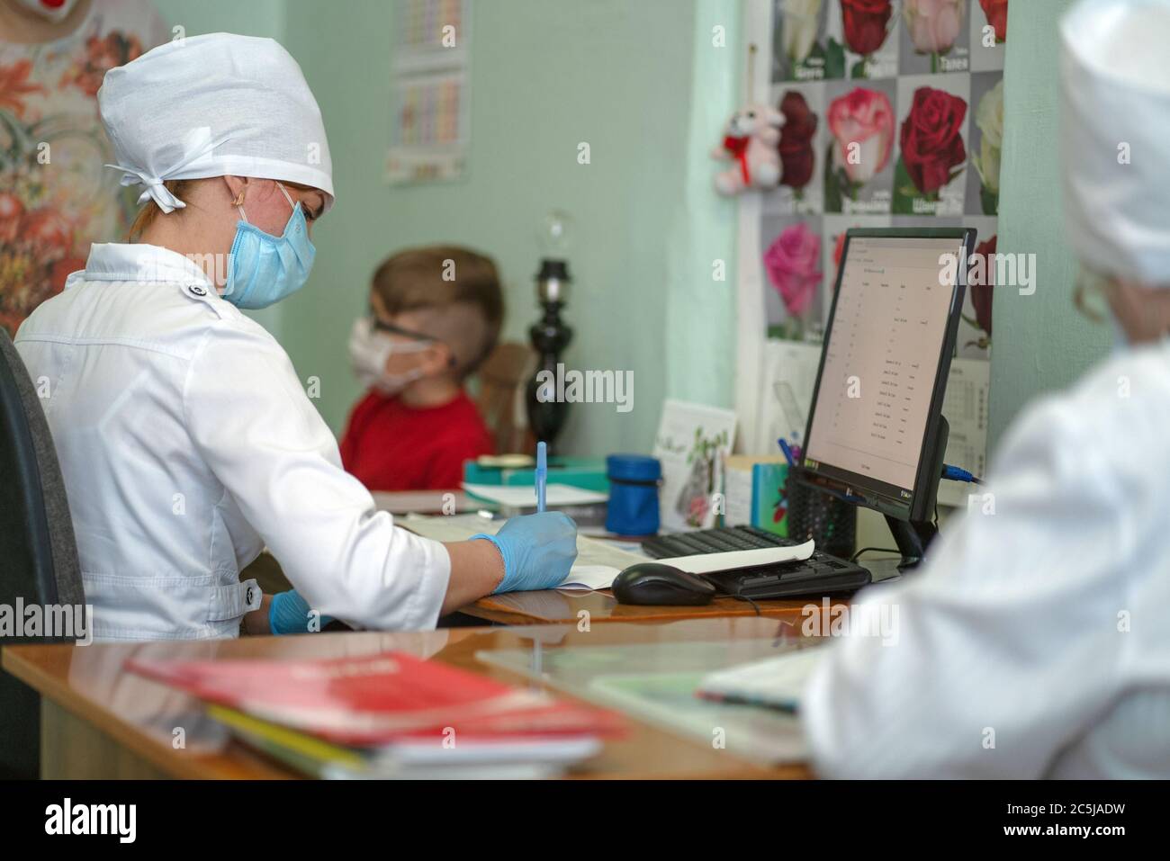 Doctor makes notes on card of little patient after examination, recording appointment and results. Masked woman and boy due to virus outbreak and Stock Photo