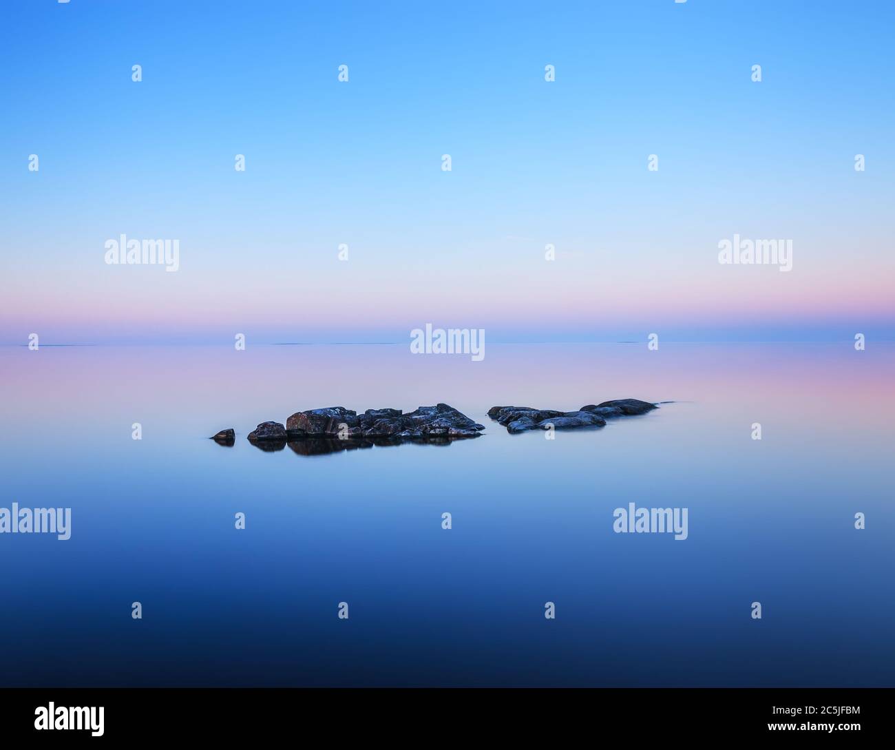 Tranquil minimalist landscape with rocks on the smooth surface of the lake with calm water with horizon with clear blue sky in twilight. Stock Photo