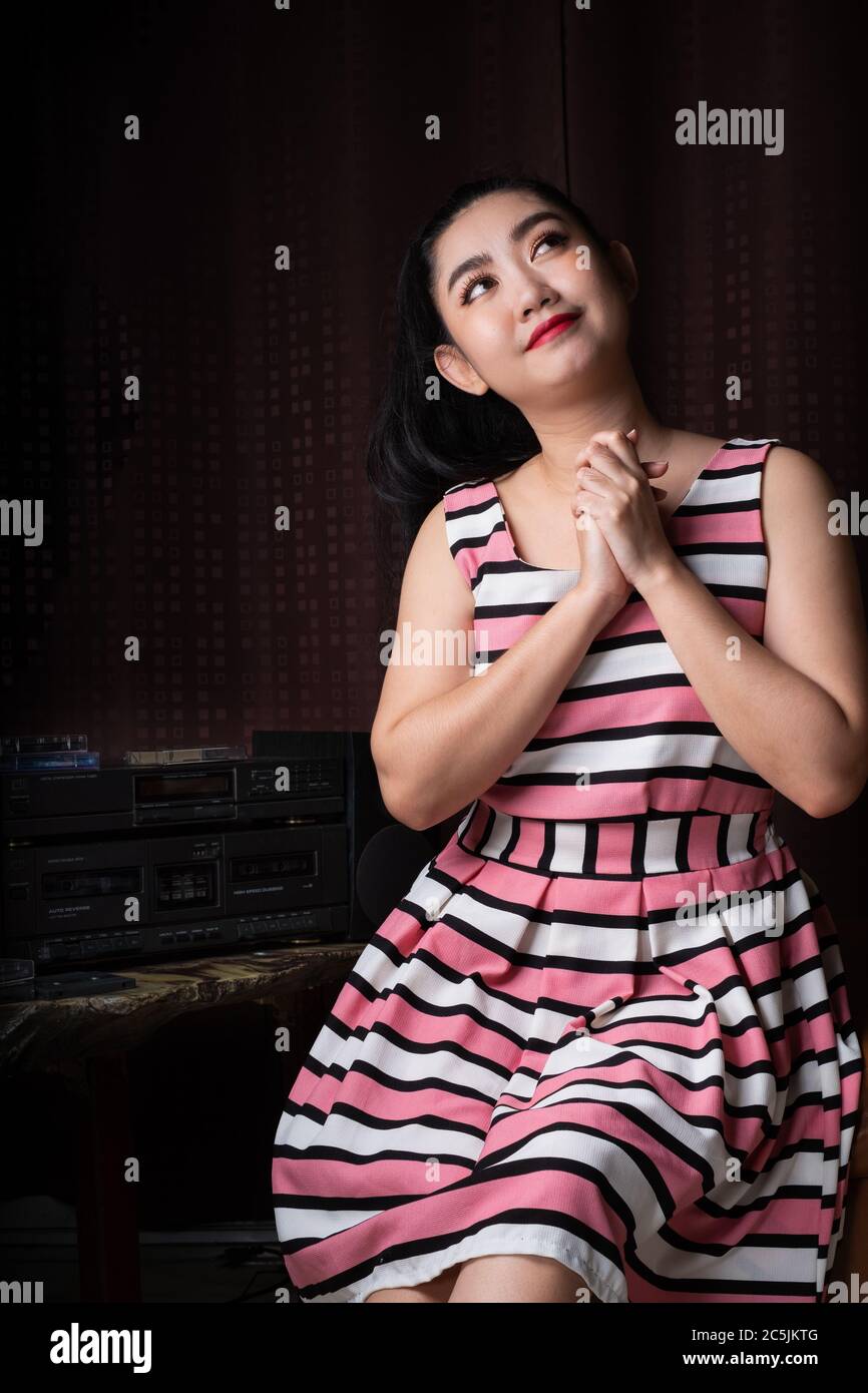 Portrait young Asia woman lolls on a black leather sofa in retro fashion pink dress listening to music from stereo cassette tape player is retro techn Stock Photo