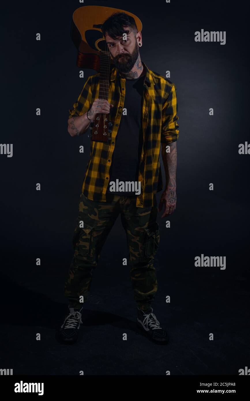 Tattooed man with a beard wearing a tartan yellow black shirt is posing holding his guitar on his sholder Stock Photo