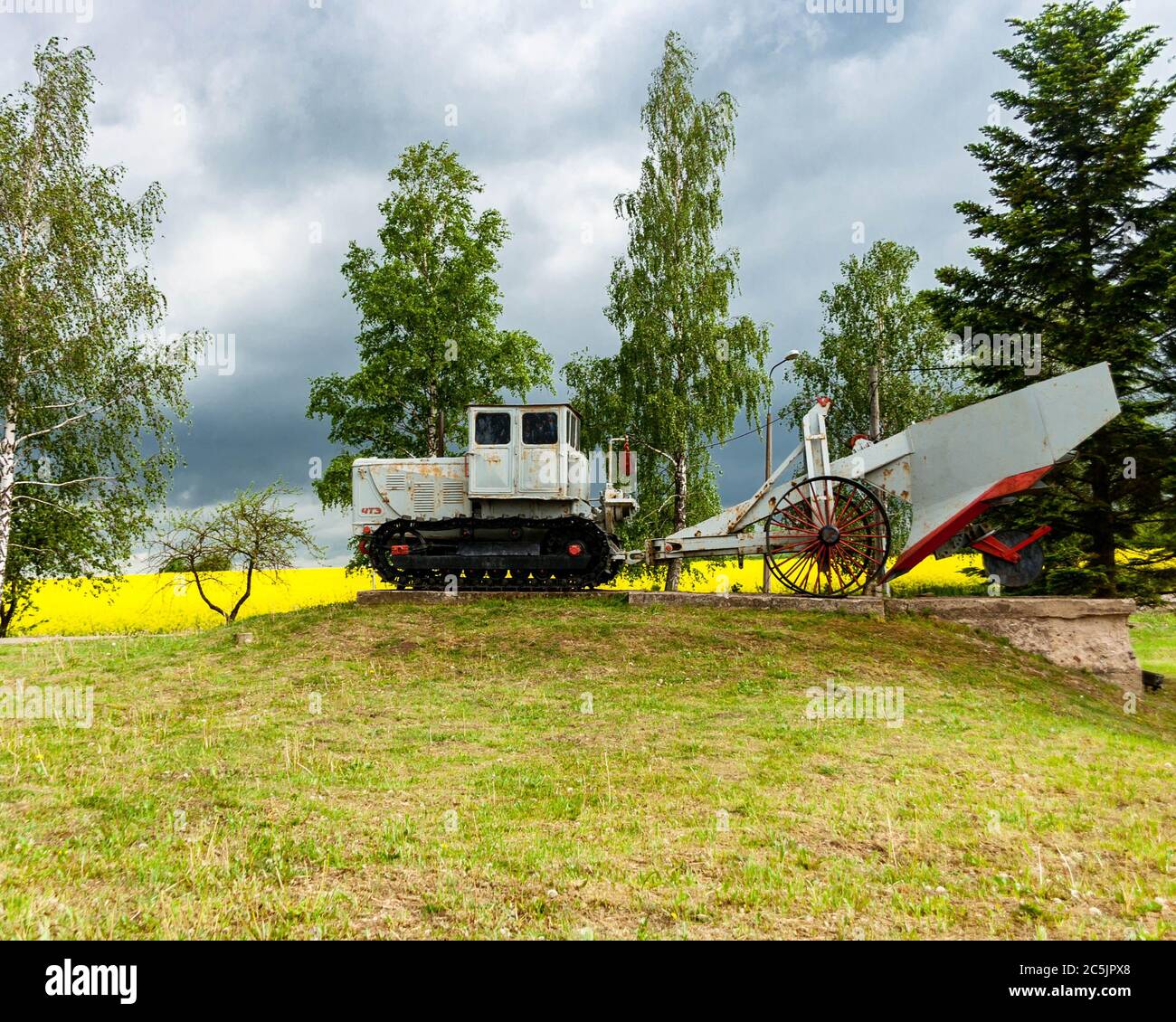 Old, rusty Soviet caterpillar tractor made in Chelyabinsk Tractor Plant with attached agriculture machinery displayed near rapeseed field. Stock Photo