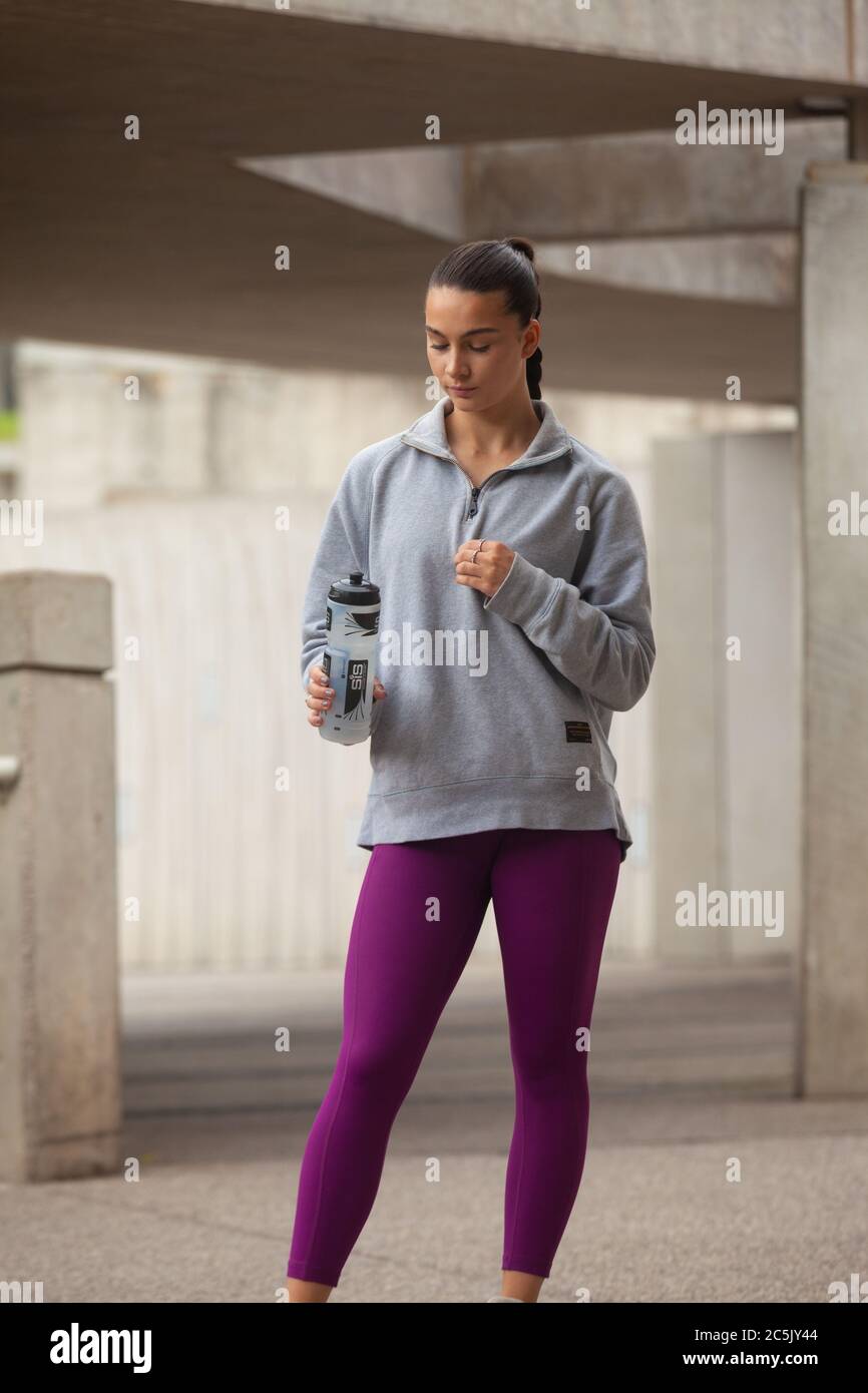 A woman standing wearing purple leggings, grey sweatshirt  and holding a drinks bottle Stock Photo