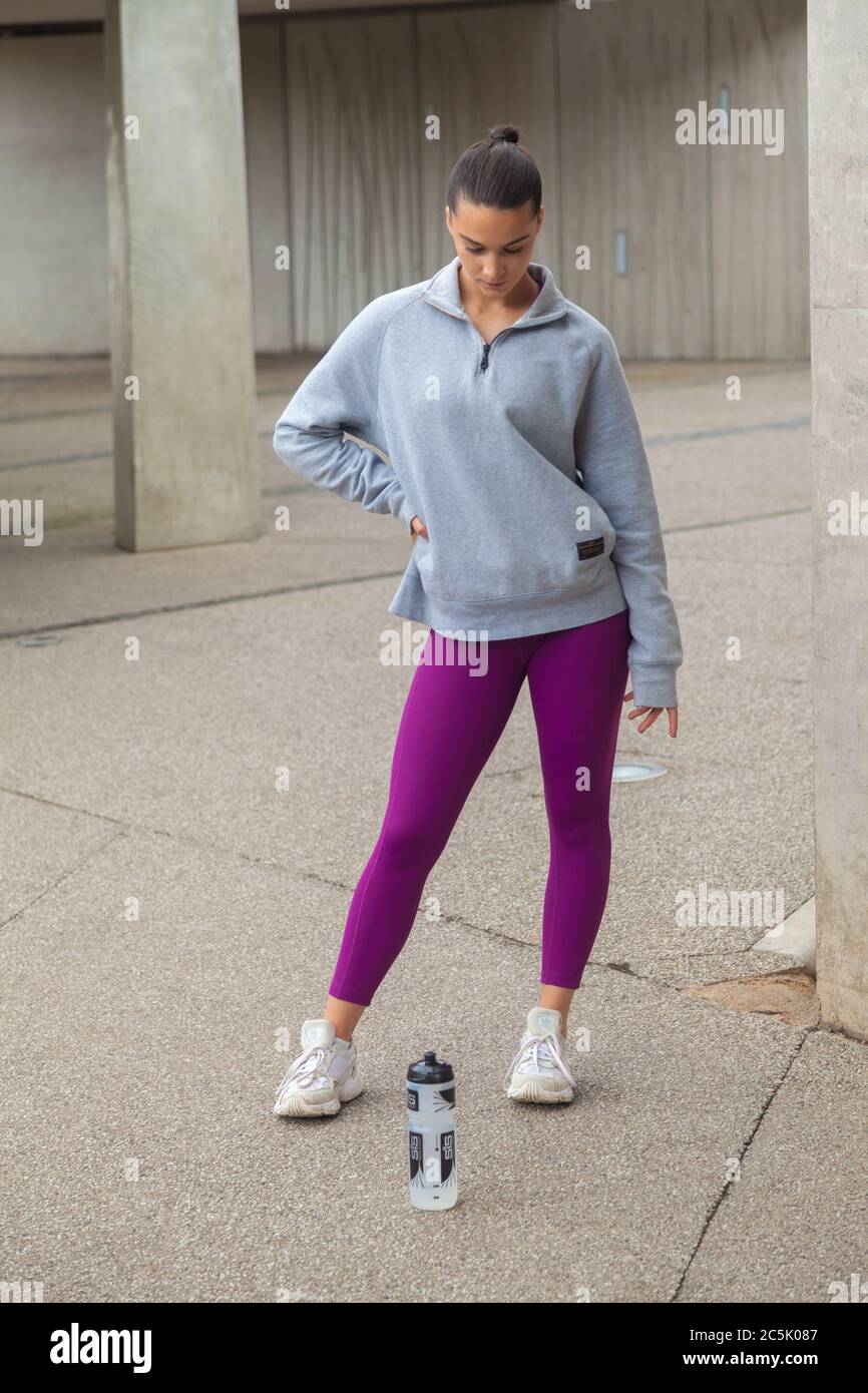 A woman standing wearing purple leggings, grey sweatshirt  and looking at a drinks bottle on the ground. Stock Photo
