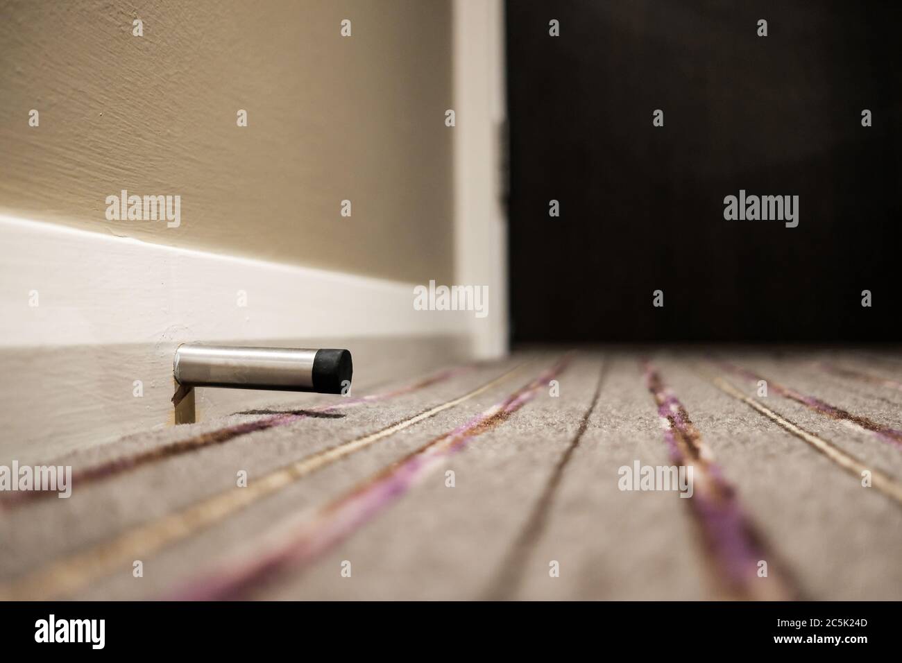 Shallow focus, from ground level of a metal door stop seen attached to a  skirting board in a modern hotel room. A nearby hotel room door is seen  also Stock Photo -, image size:1300x956