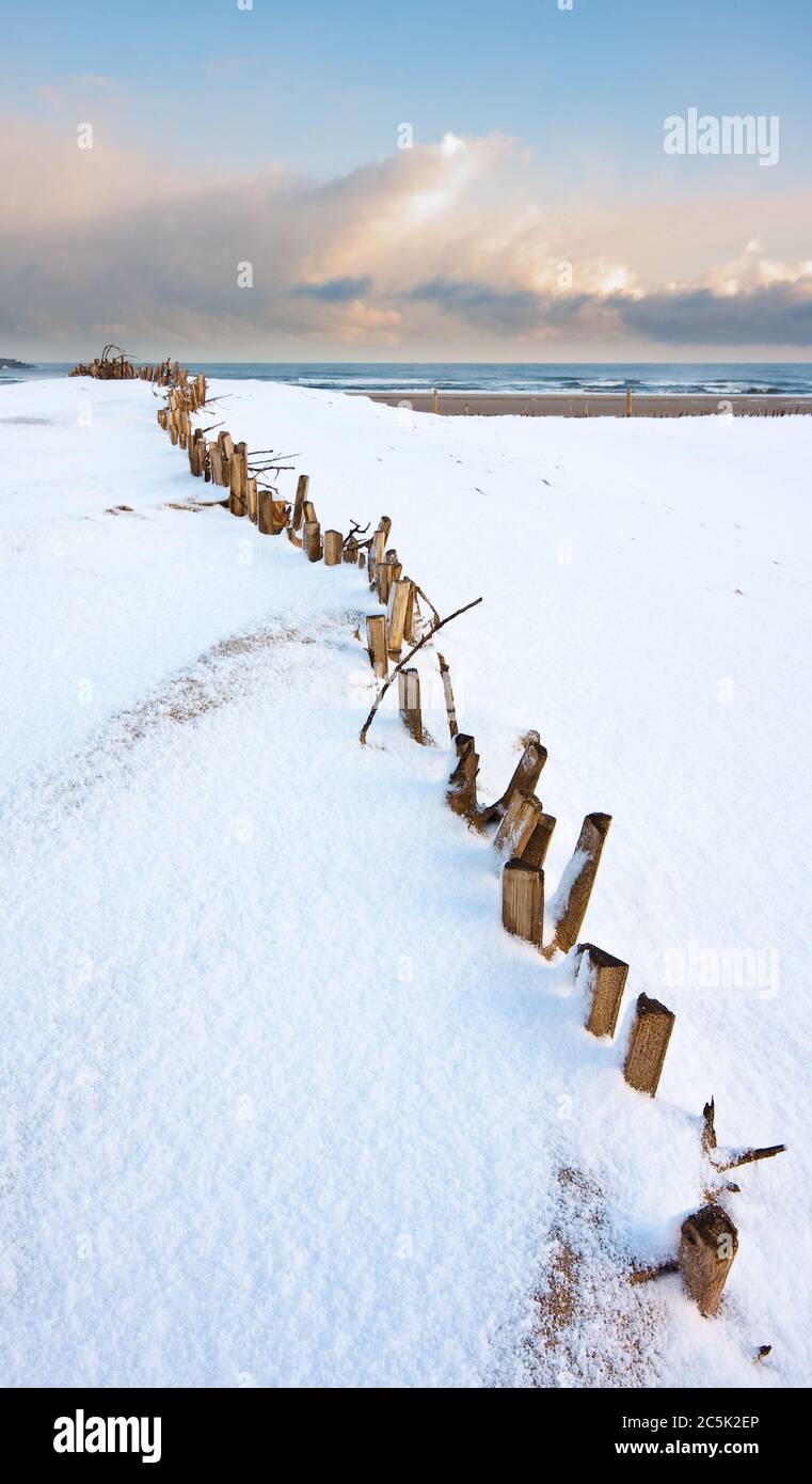 Looking over the snow covered sand dunes which have buried the fence in the foreground towards the storm clouds on the ocean towards the horizon. Stock Photo