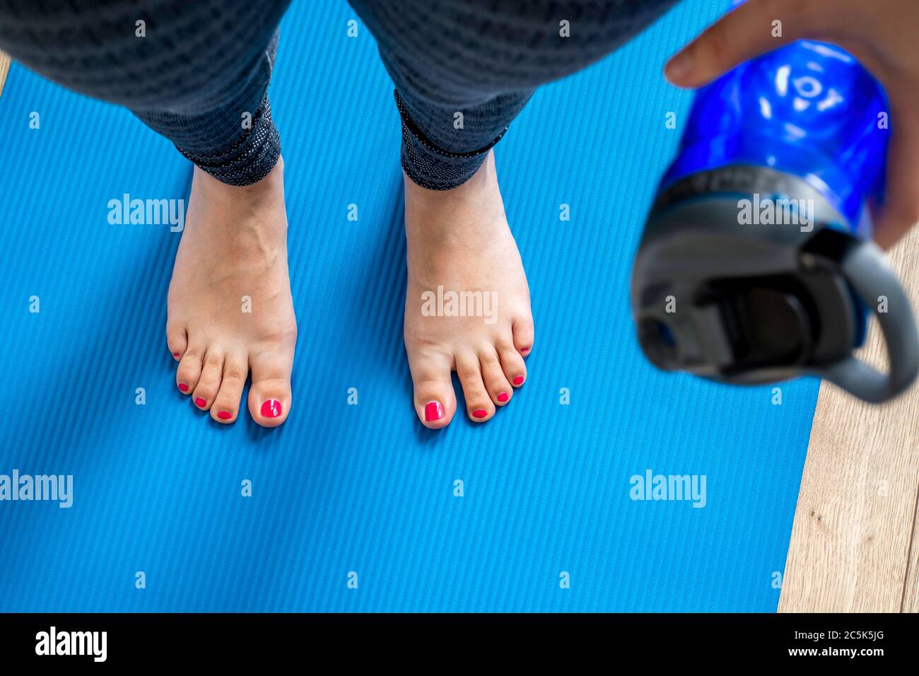 Woman practicing yoga, holding a bottle water. Feet with pink pedicure of woman on blue mat, first person top view. Concept of indoor workout Stock Photo