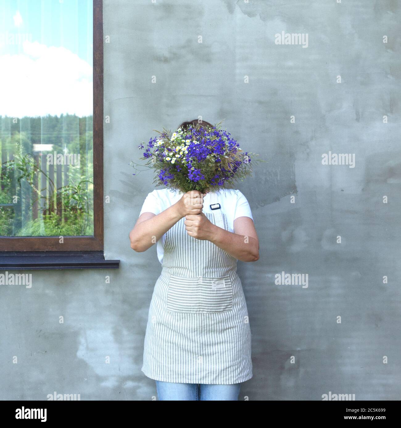 The girl in an apron hides her face behind a bouquet of meadow flowers. The window reflects the sky and the fence. F Stock Photo