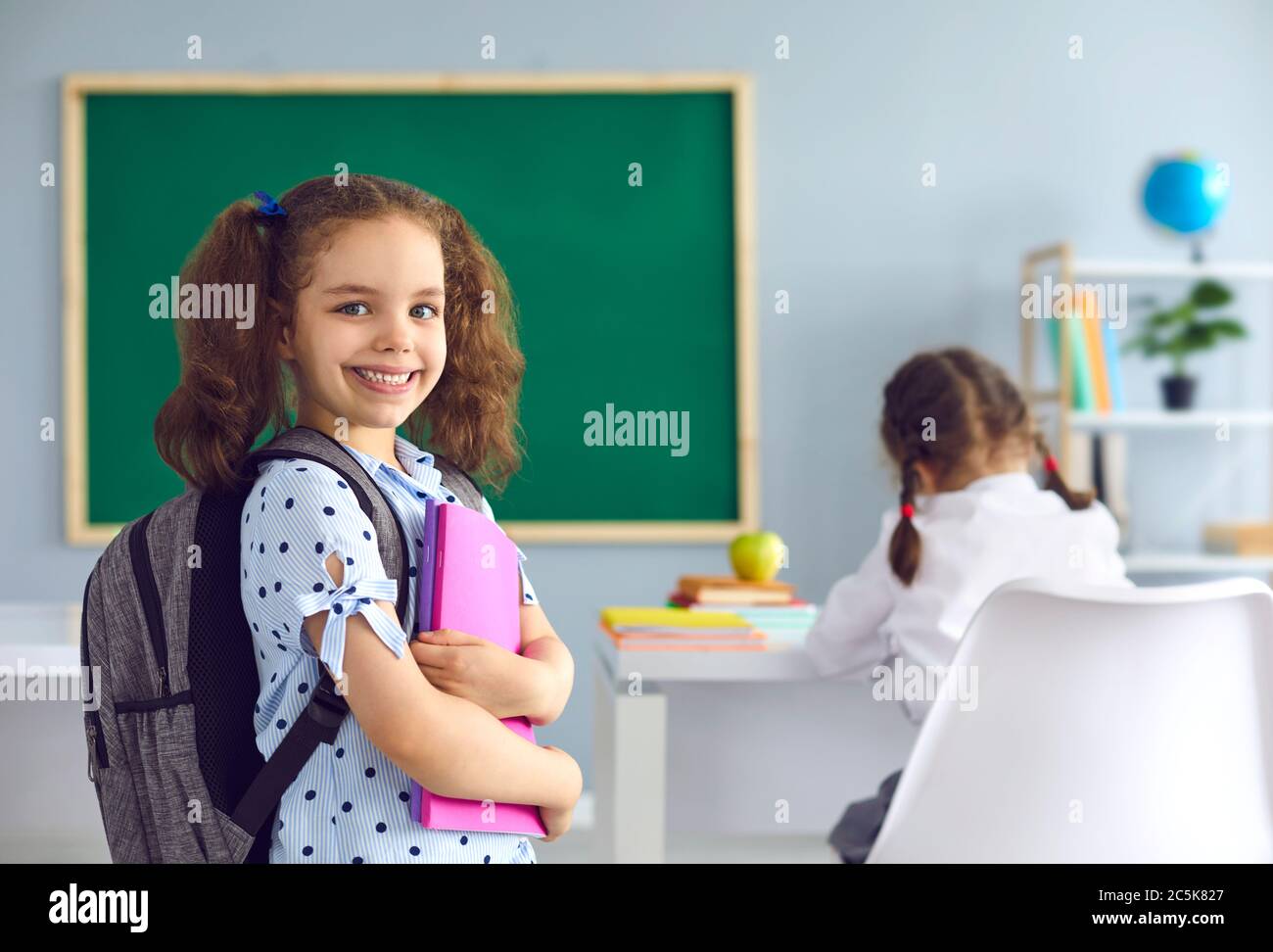 Back to school. Smiling schoolgirl with a book in her hand looks at the camera while standing in class. Stock Photo