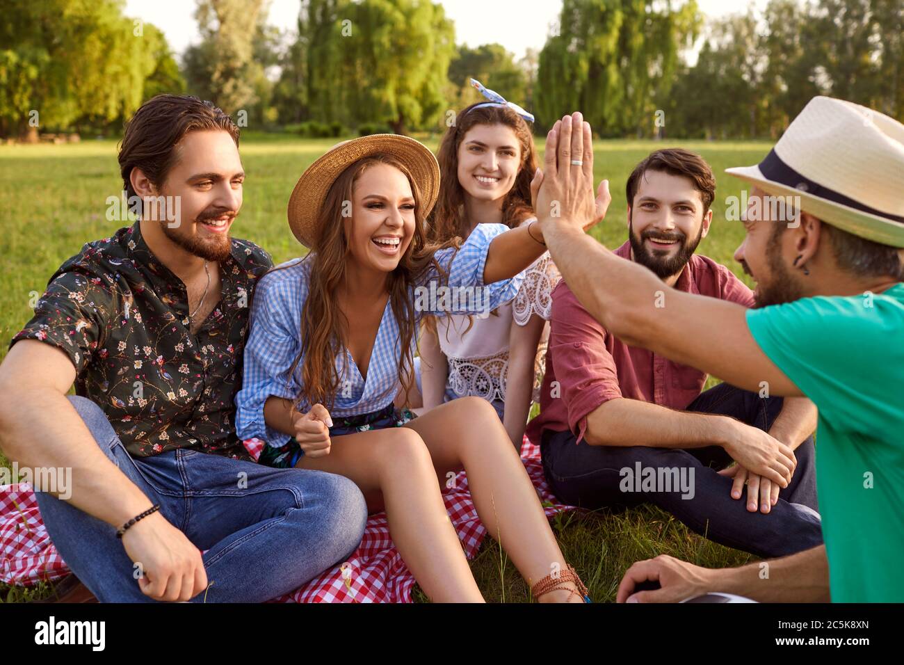 Group of friends sitting on grass, talking and high fiving in countryside. Joyful people enjoying time together outdoor in park Stock Photo