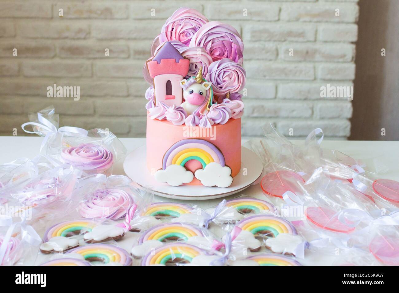 Pink birthday cake with fondant unicorn, rainbow and meringue clouds. Candy bar with cake, cookies and lollipops. Stock Photo
