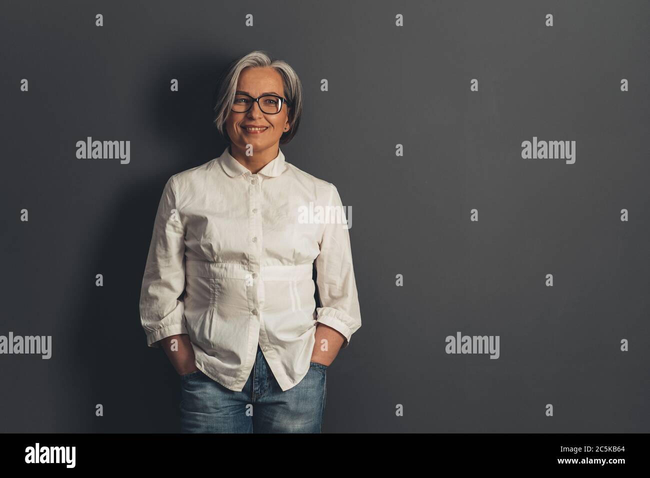 Confident mature woman toothy smiling while standing hands in pockets. Happy senior woman cut out on gray background with copy space at right side Stock Photo