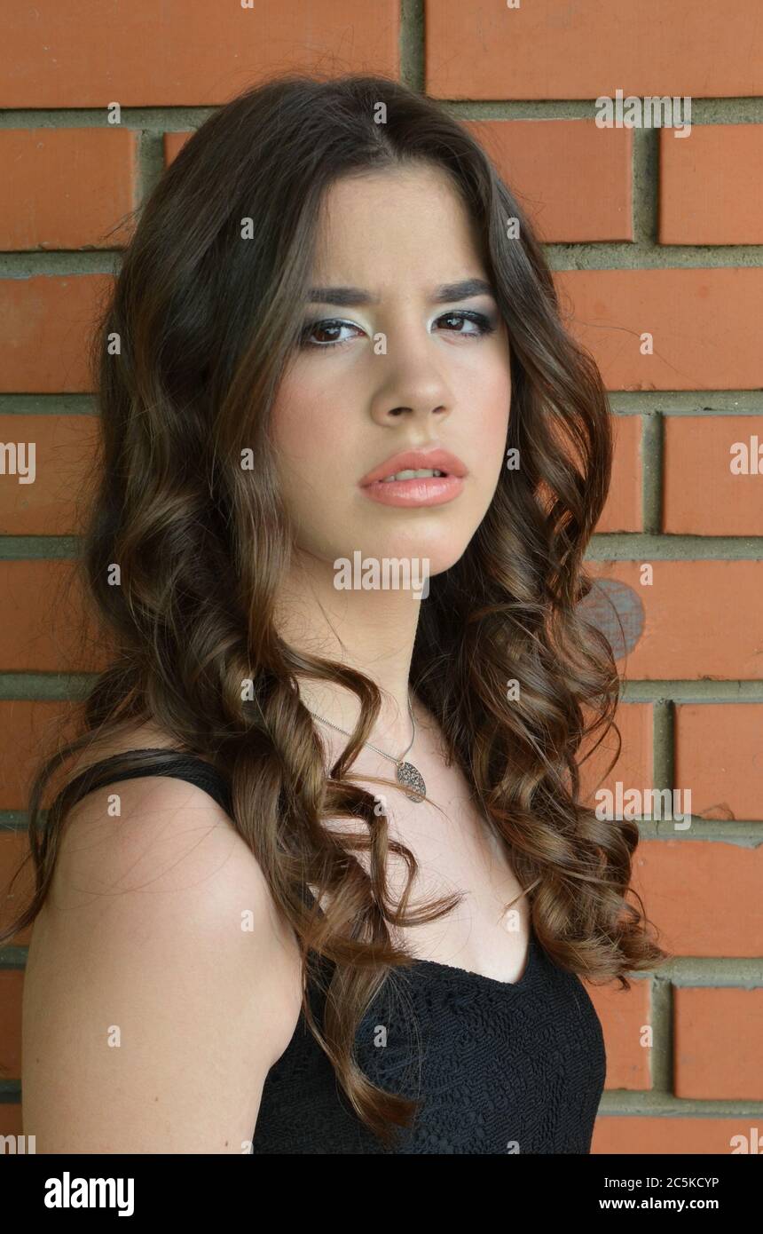 Portrait of cute brunette teenage girl in black dress against red brick wall background Stock Photo