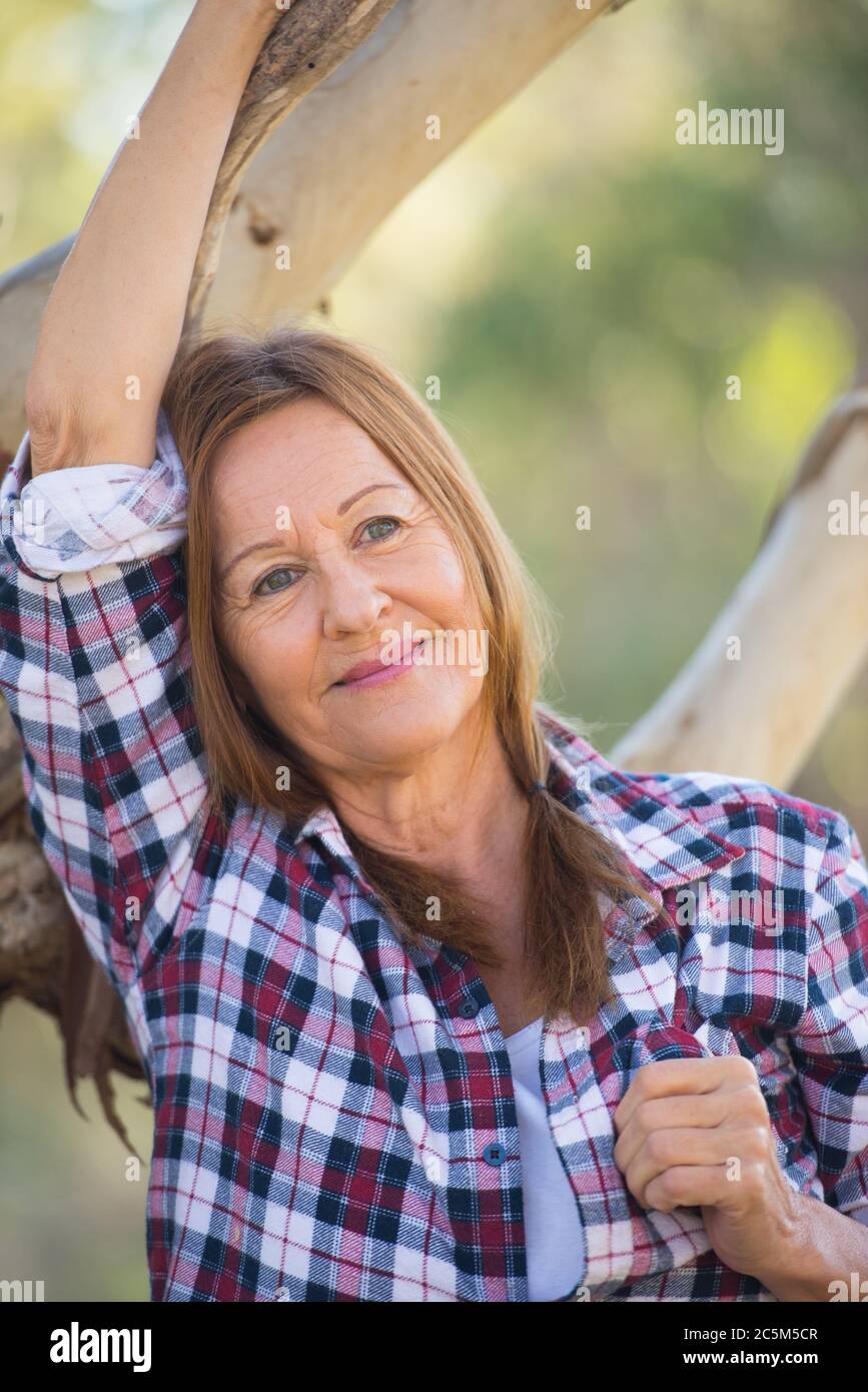Portrait attractive mature woman in rural country, wearing plaid shirt, posing relaxed thoughtful with happy smile against Australian outback bush bac Stock Photo