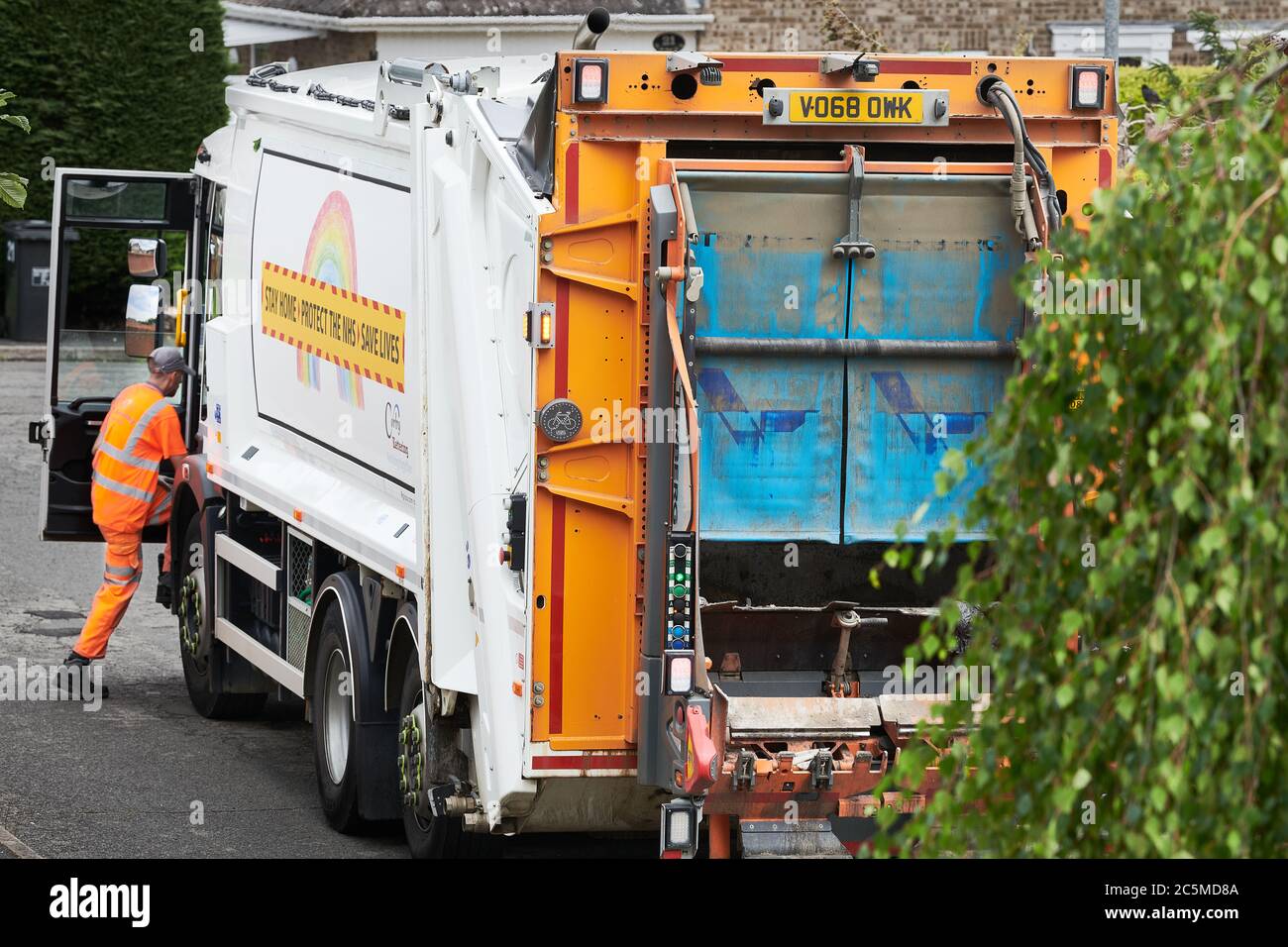 A recycling collector, in high visibility orange clothing, enters a refuse collection lorry after he has emptied a bin of rubbish into the back of it. Stock Photo