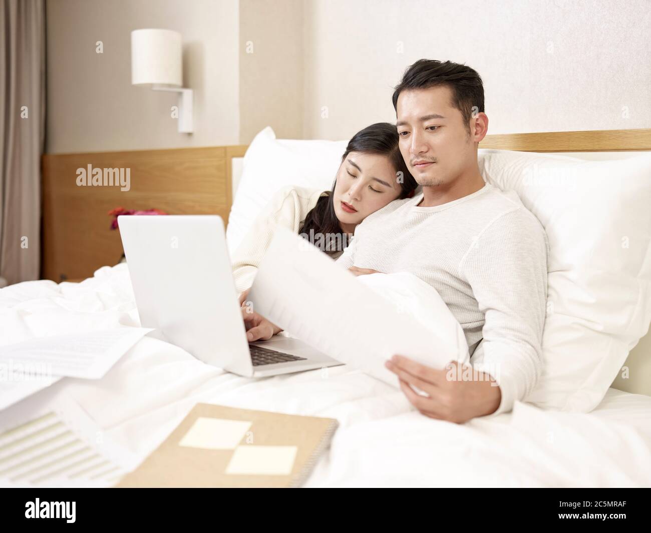 young asian business man sitting in bed working using laptop computer while wife falling asleep by his side Stock Photo