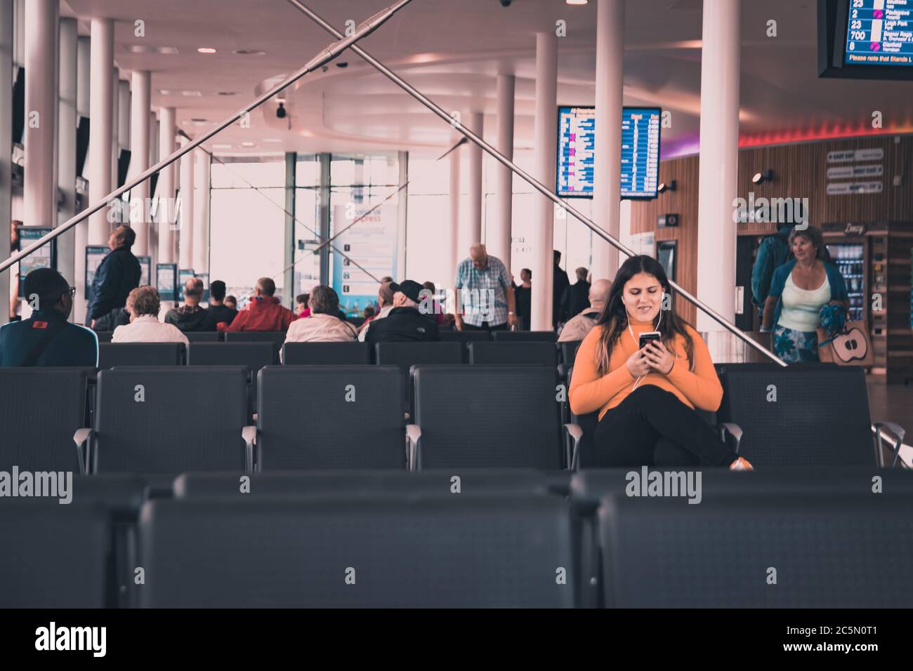 a young woman looking at her phone with headphones in smiling while seated waiting a a terminal for transport to arrive Stock Photo