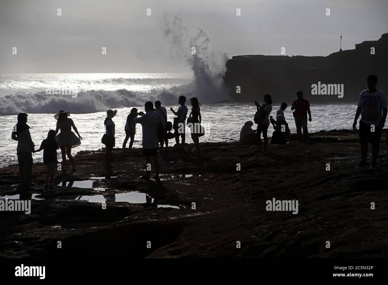 Tourists silhouetted as they have leisure time on a rocky beach with large wave smashing on cliff in the background, in Tanah Lot, Bali, Indonesia. Stock Photo
