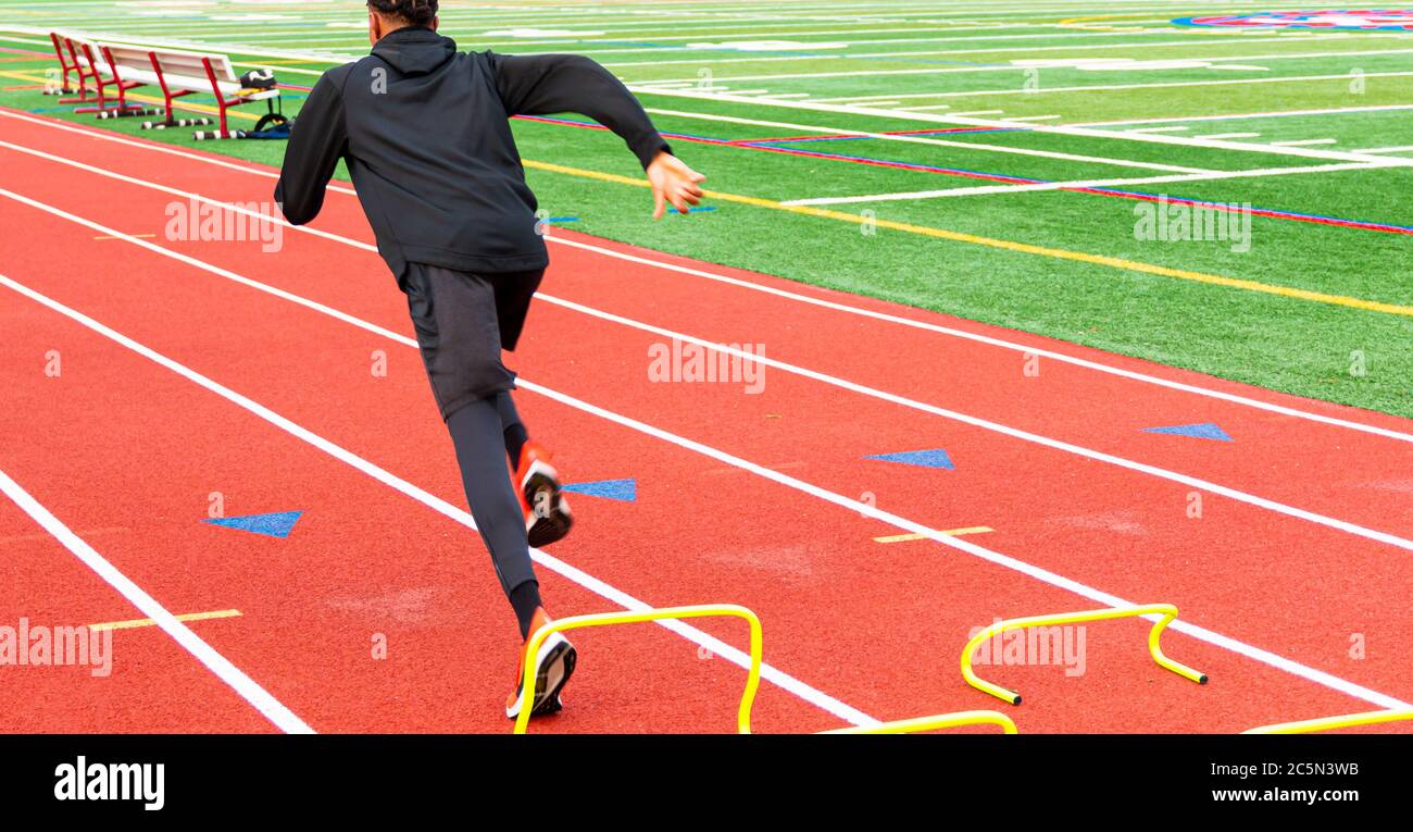 Rear view of a high school track and field sprinter running over yellow mini banana hurdles on a track during practice, Stock Photo
